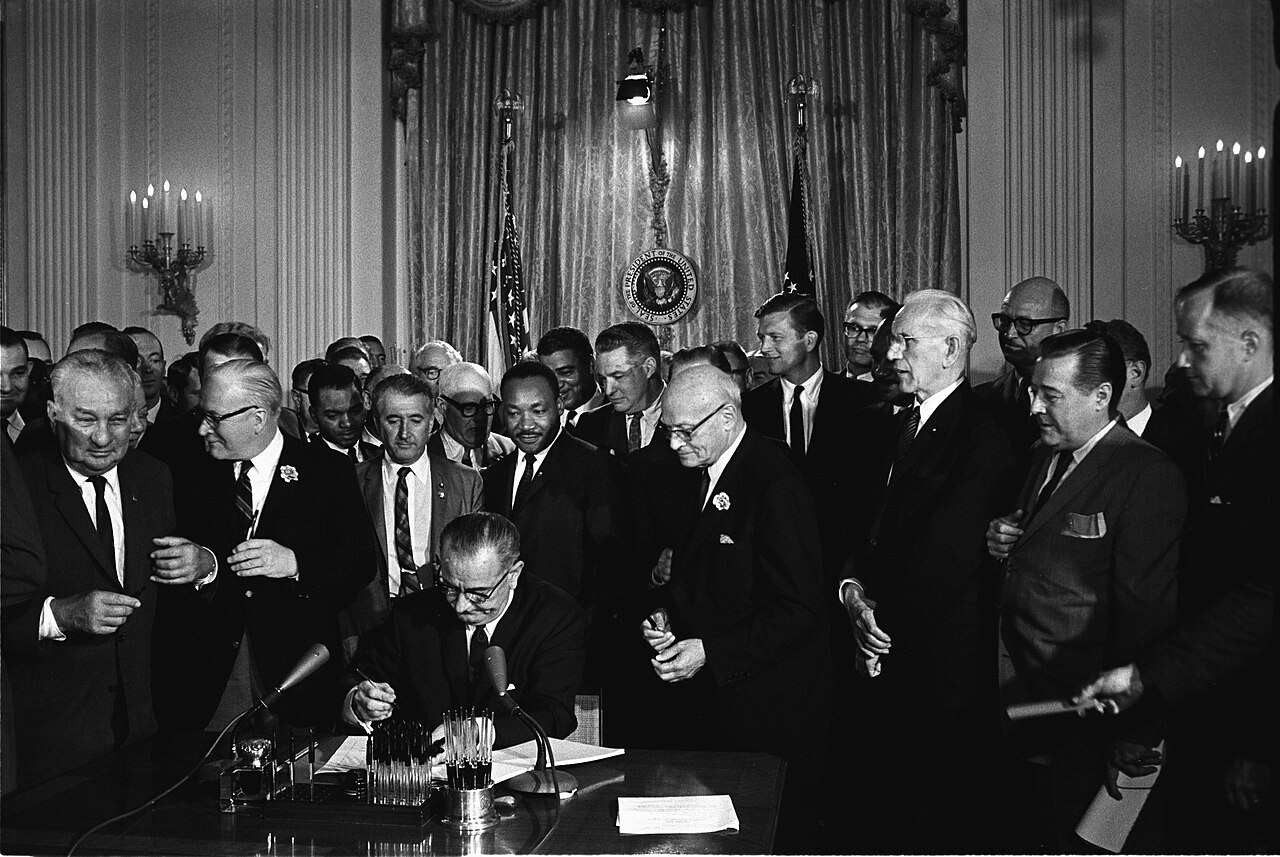 President Lyndon B. Johnson signs the Civil Rights Act of 1964, with Dr. Martin Luther King Jr. and others standing behind him.