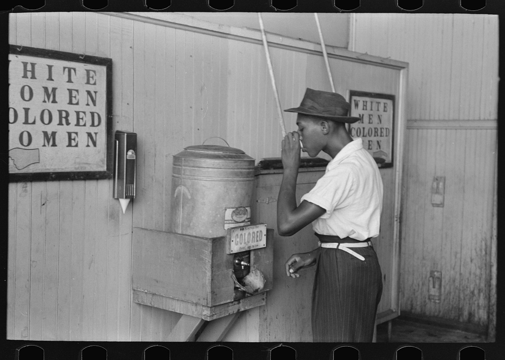 A Black man drinks from a 'Colored' water cooler at a streetcar terminal in Oklahoma City, 1939.